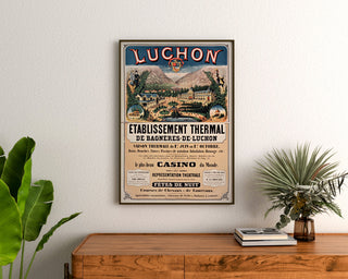 Manifesto vintage Bagnères-de-Luchon, terme, incorniciato sopra una cassettiera in legno, tra delle piante.