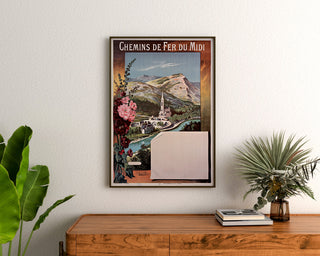 Manifesto vintage Lourdes, ferrovie del Midi, incorniciato sopra una cassettiera in legno, tra piante.